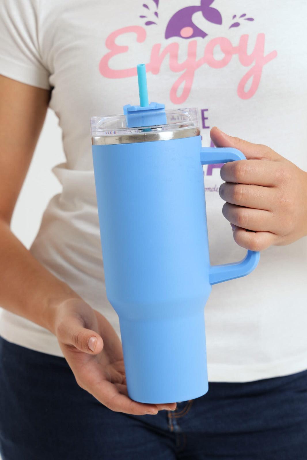 Person holding a blue insulated tumbler with a straw lid, wearing a white shirt with 'Enjoy' text.